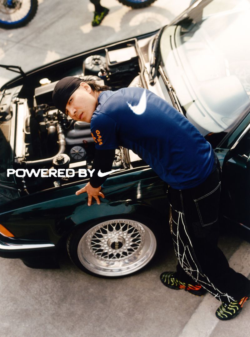 Man in Nike jersey leans on a dark green car with the hood up, showing the engine; lower part of motorbike wheel visible in background.