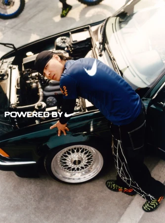 Man in Nike jersey leans on a dark green car with the hood up, showing the engine; lower part of motorbike wheel visible in background.