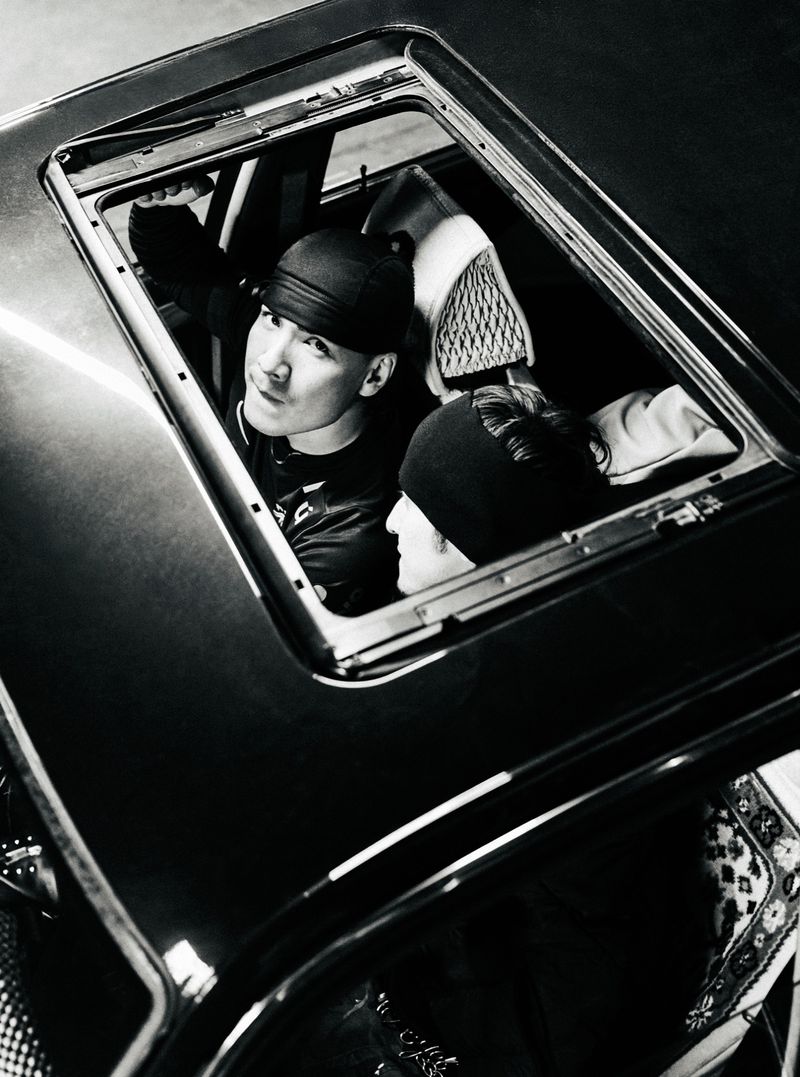 Two people, wearing beanies, are seen in a monochrome shot looking up through an open sunroof of a black vehicle.