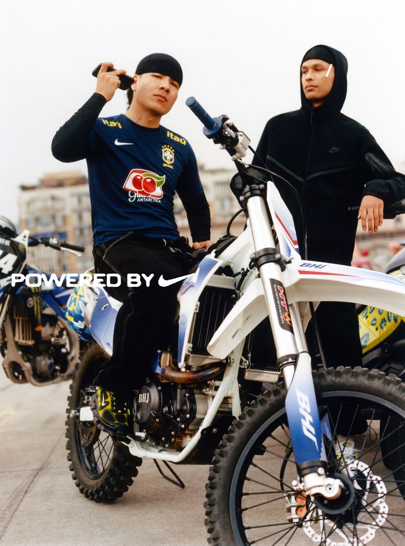 Two men pose with dirt bikes on a city street, one seated on a white bike wearing a Brazil soccer jersey and a do-rag. 