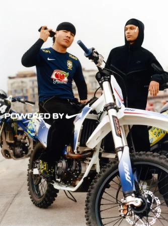 Two men pose with dirt bikes on a city street, one seated on a white bike wearing a Brazil soccer jersey and a do-rag. 