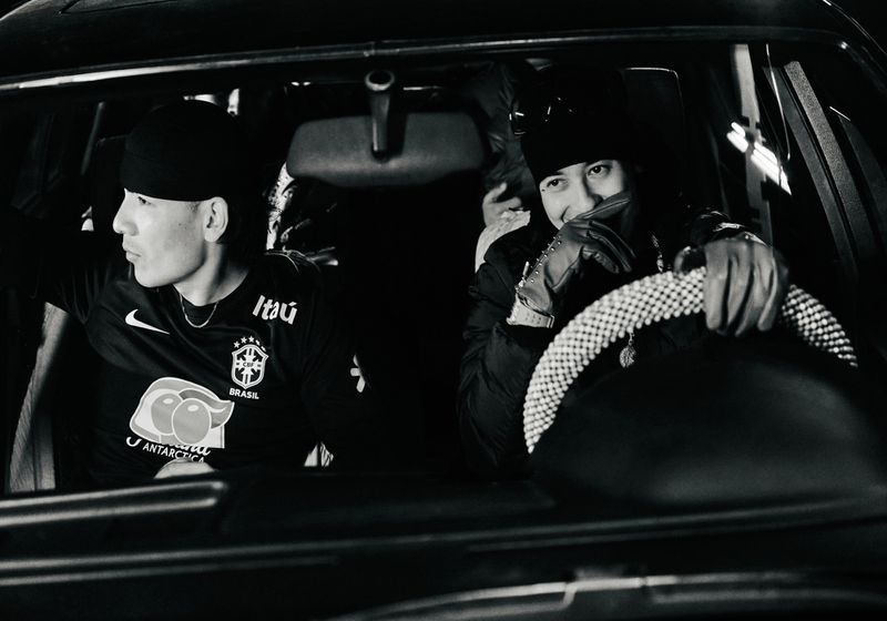 Two stylish men sit inside a car wearing beanies, one driving with a jeweled steering wheel in a monochrome, low-light interior.