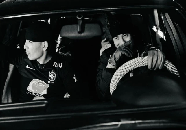 Two stylish men sit inside a car wearing beanies, one driving with a jeweled steering wheel in a monochrome, low-light interior.