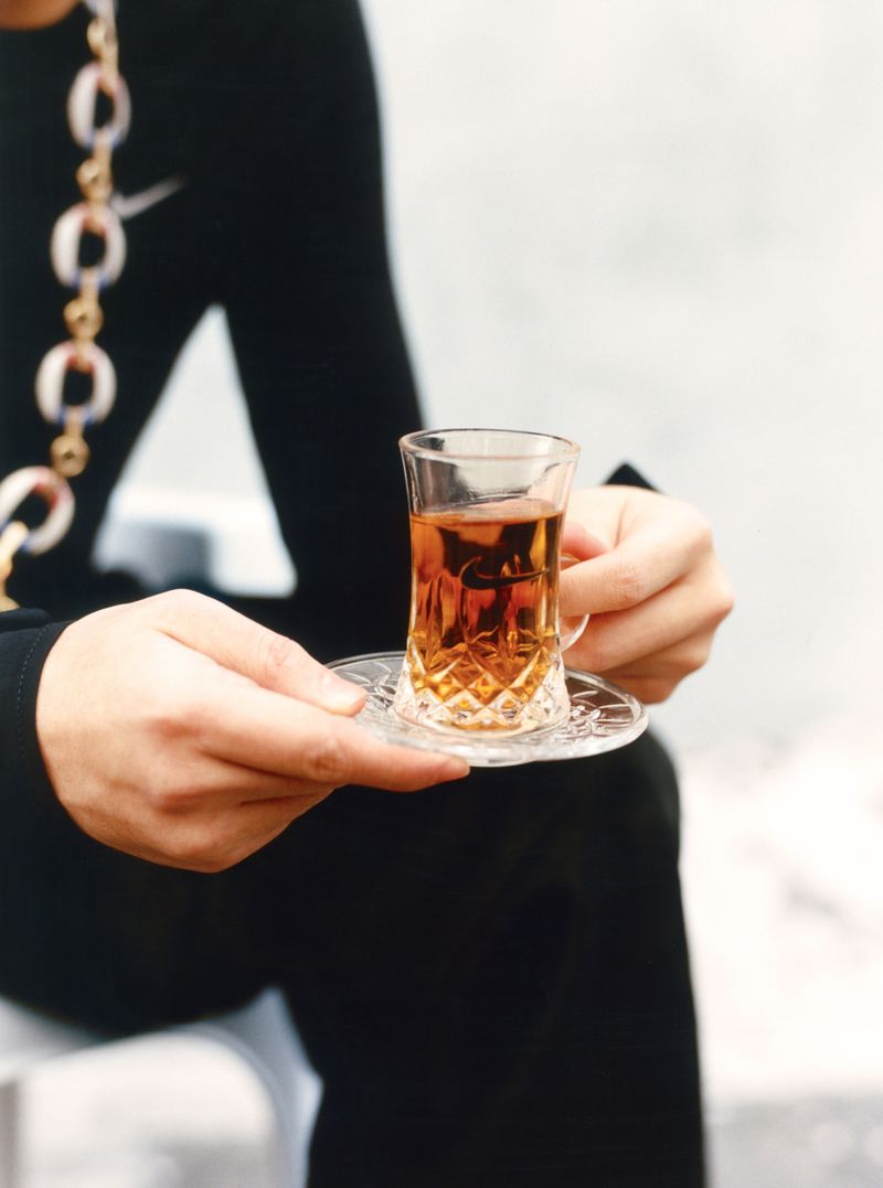 Person holding a glass of tea on a saucer, wearing a black top with a gold chain in an indoor setting with neutral background.