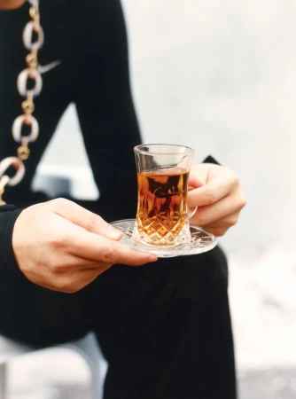 Person holding a glass of tea on a saucer, wearing a black top with a gold chain in an indoor setting with neutral background.
