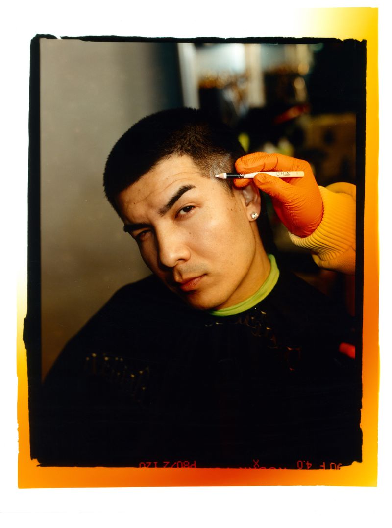Young man getting eyebrow styling by gloved stylist using pen. Hair and beauty concept shot with black styling cape indoors.