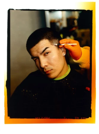 Young man getting eyebrow styling by gloved stylist using pen. Hair and beauty concept shot with black styling cape indoors.