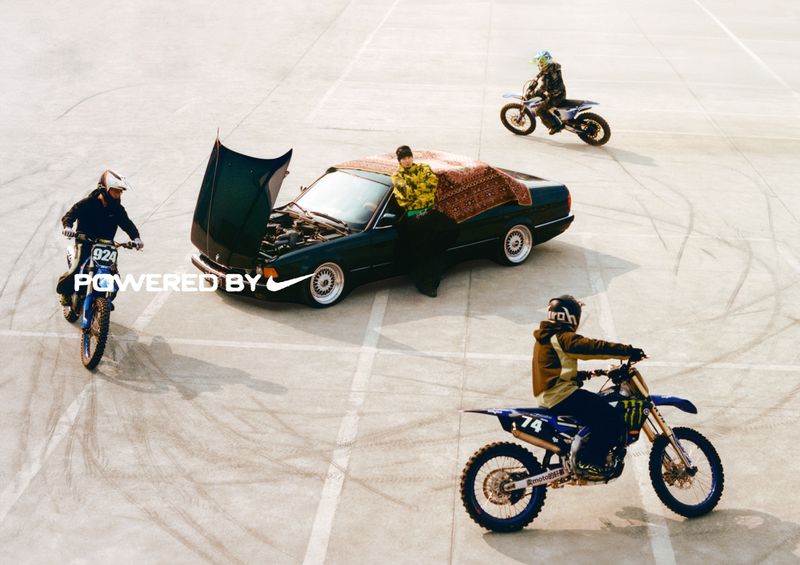 A dark green car with its hood open is parked on a paved surface as dirt bikers ride in a circle, creating tire track marks.