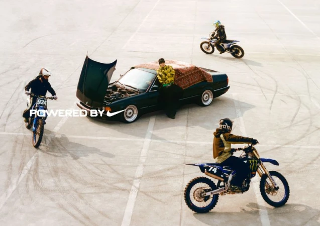 A dark green car with its hood open is parked on a paved surface as dirt bikers ride in a circle, creating tire track marks.