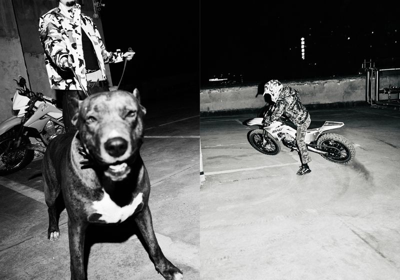 Monochrome image showcases a large dog and a man with a motorcycle, with a second man performing a motorcycle stunt, captured at night.
