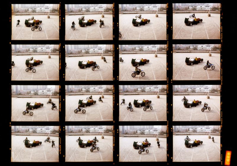 Contact sheet of motorcycle stunt driving over car on paved lot with city buildings in background. Motorbikes jumping and driving.