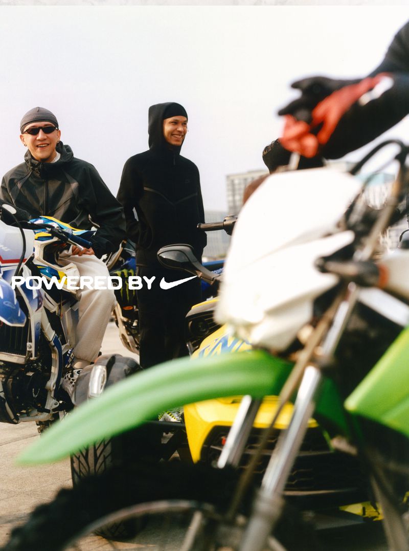 Two men on motocross bikes outdoors, one wearing a beanie and jacket, the other a hooded sweatshirt. Nike logo and text overlay.