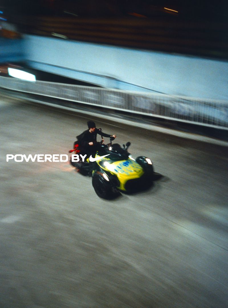 A person rides a yellow and black three-wheeled vehicle on a gray road under a gray bridge in an action-filled outdoor shot.