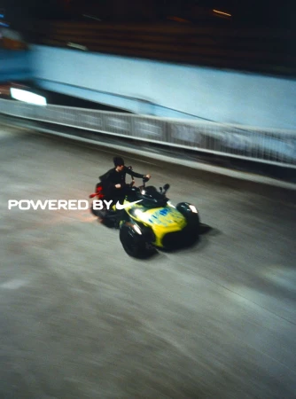 A person rides a yellow and black three-wheeled vehicle on a gray road under a gray bridge in an action-filled outdoor shot.