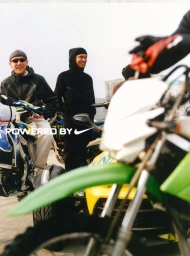 Two smiling men in black jackets with motorcycles outdoors under an overcast sky for a Nike campaign photographed by Hailun Ma.