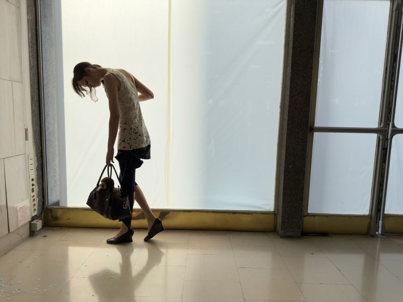 A slender woman in a lace top and dark shorts carries a large handbag in a brightly lit indoor space with large windows.