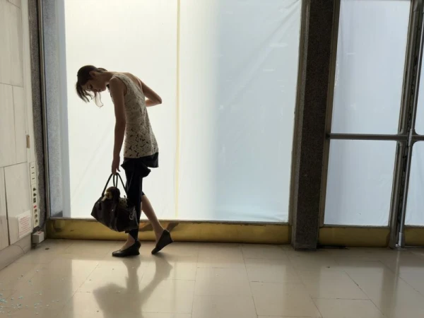 A slender woman in a lace top and dark shorts carries a large handbag in a brightly lit indoor space with large windows.