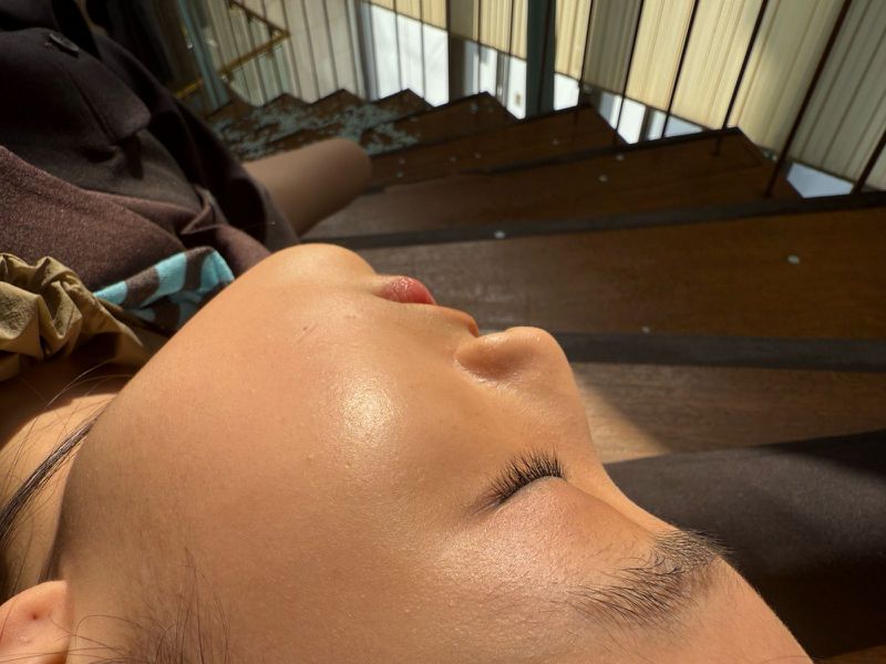 Close-up of an adult female face with natural makeup, eyes closed, lying down outdoors in soft sunlight against a wooden background.