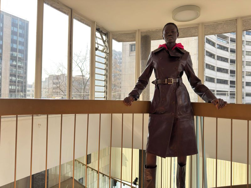 Woman in brown leather trench coat stands in modern interior with geometric railings and floor-to-ceiling windows overlooking cityscape.
