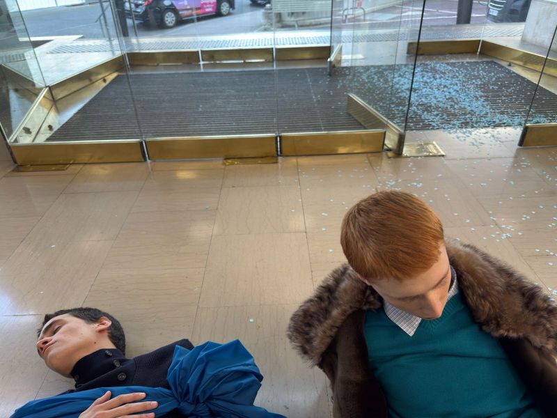 Two men lie on a marble floor near a shattered glass entrance. One wears a fur coat, the other a blue outfit, indoor setting.