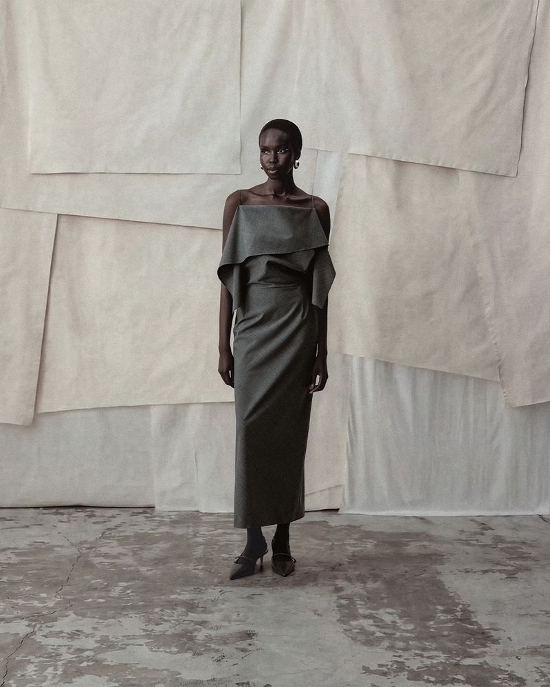 Elegant African woman poses in a stylish draped gray dress against a background of textured white panels, showcasing a refined fashion aesthetic.