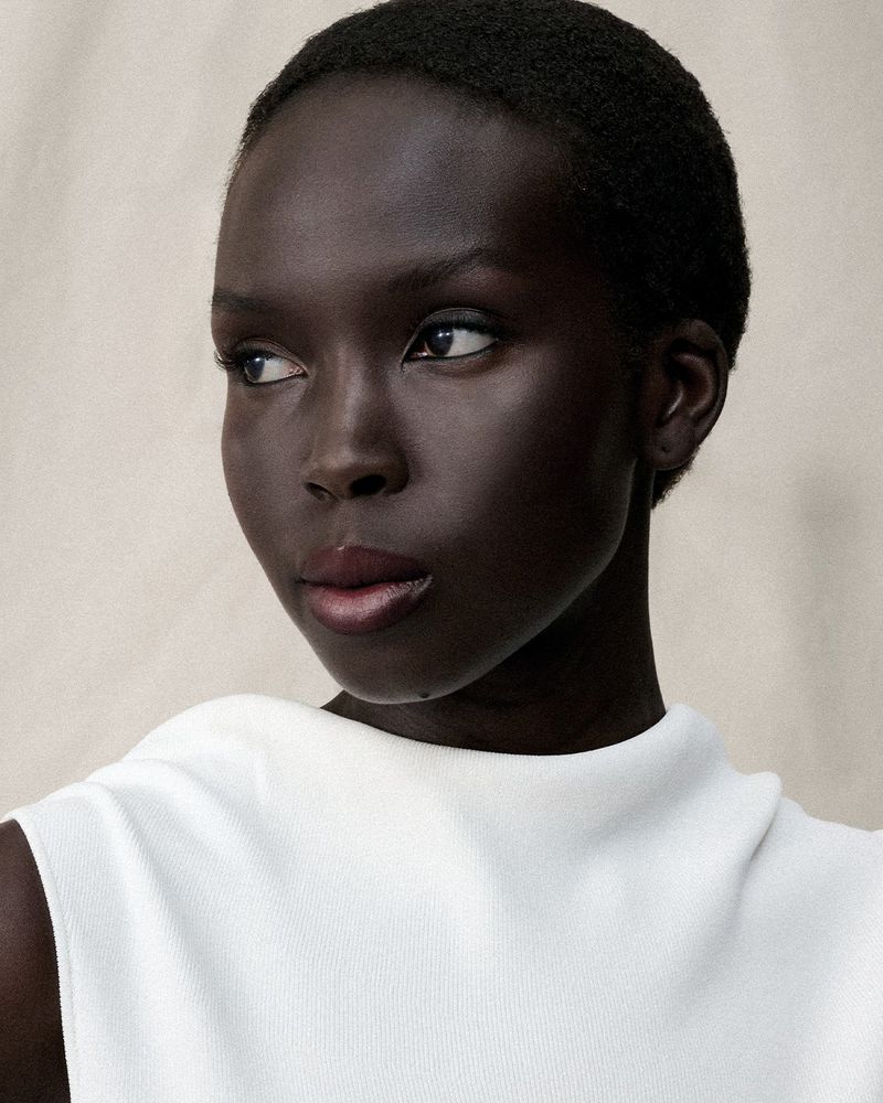 Portrait of a dark-skinned African woman with short hair wearing a white sleeveless top against a neutral background, looking left.