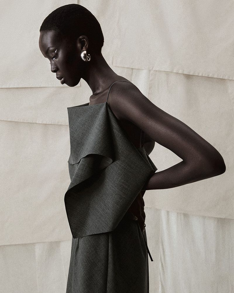 An African-American woman poses in a gray outfit with ruffled details against a neutral backdrop in a fashion forward studio shot.