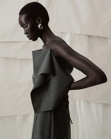 An African-American woman poses in a gray outfit with ruffled details against a neutral backdrop in a fashion forward studio shot.