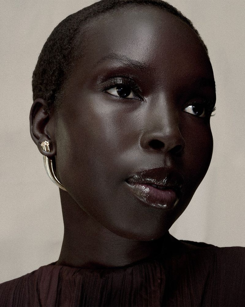 Close-up portrait of a woman with short hair, wearing gold hoop earrings, glossy lips, and a dark brown top, against a neutral background.