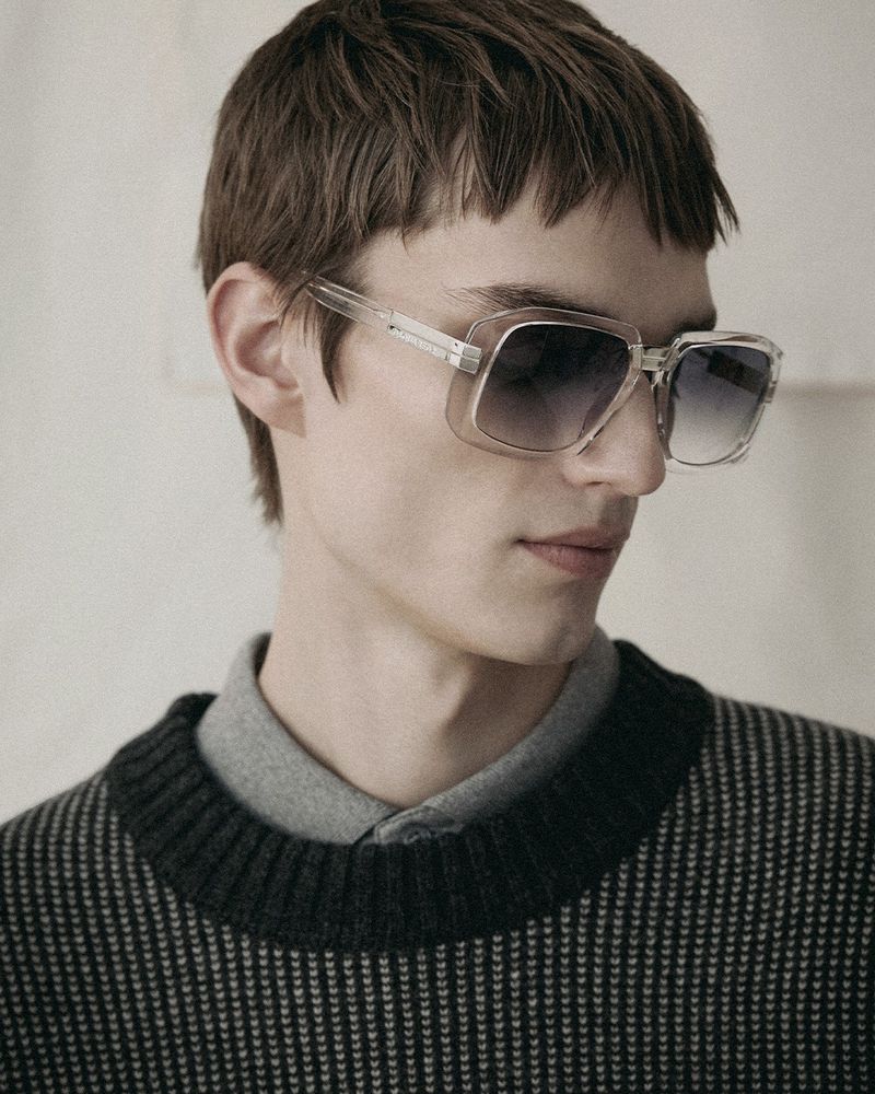 Close-up of a young adult man wearing a gray sweater and transparent square sunglasses with an ombré lens on a neutral backdrop.