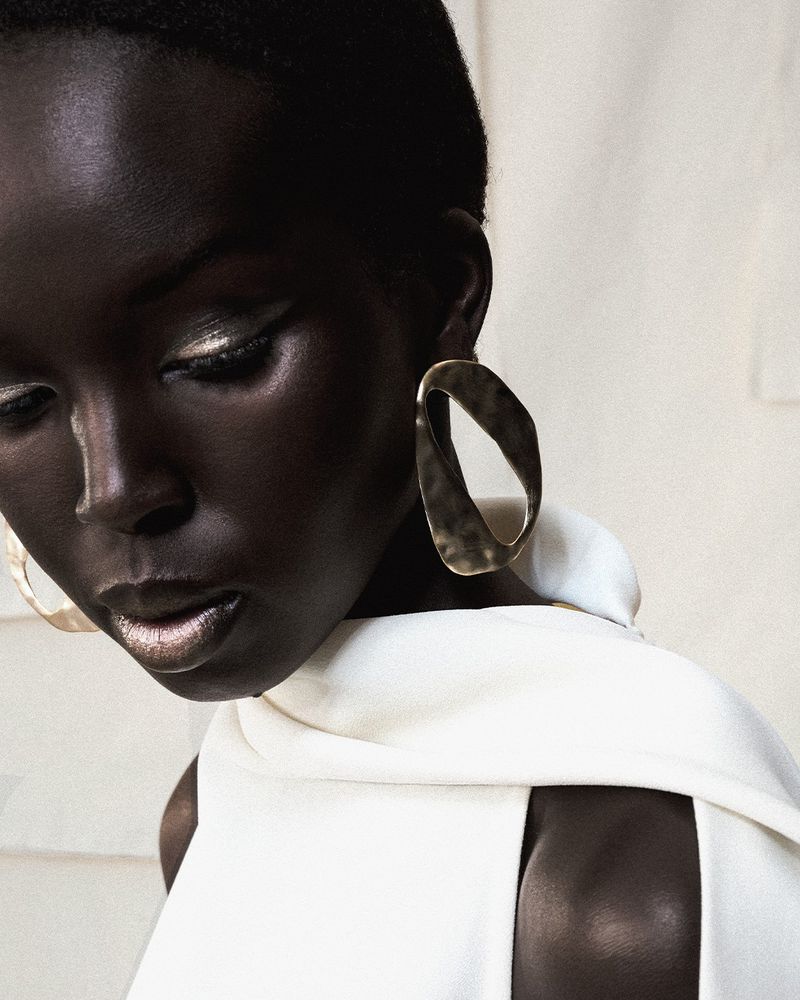 Close-up portrait of a dark-skinned woman with gold eye makeup and sculptural gold earrings, wearing a cream-colored sleeveless top.