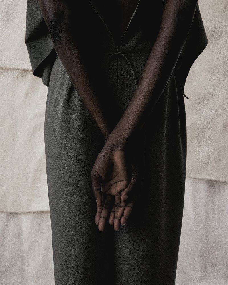 Model wearing dark grey tailored trousers with hands clasped behind back against light textured backdrop
