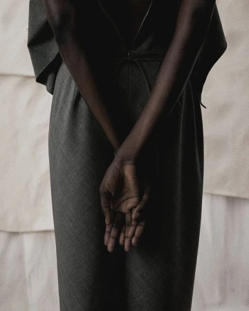 Model wearing dark grey tailored trousers with hands clasped behind back against light textured backdrop