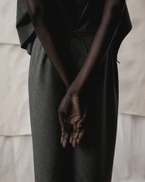 Model wearing dark grey tailored trousers with hands clasped behind back against light textured backdrop