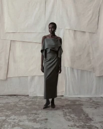 Model standing against a textured paper backdrop wearing a dark grey strapless dress and heels