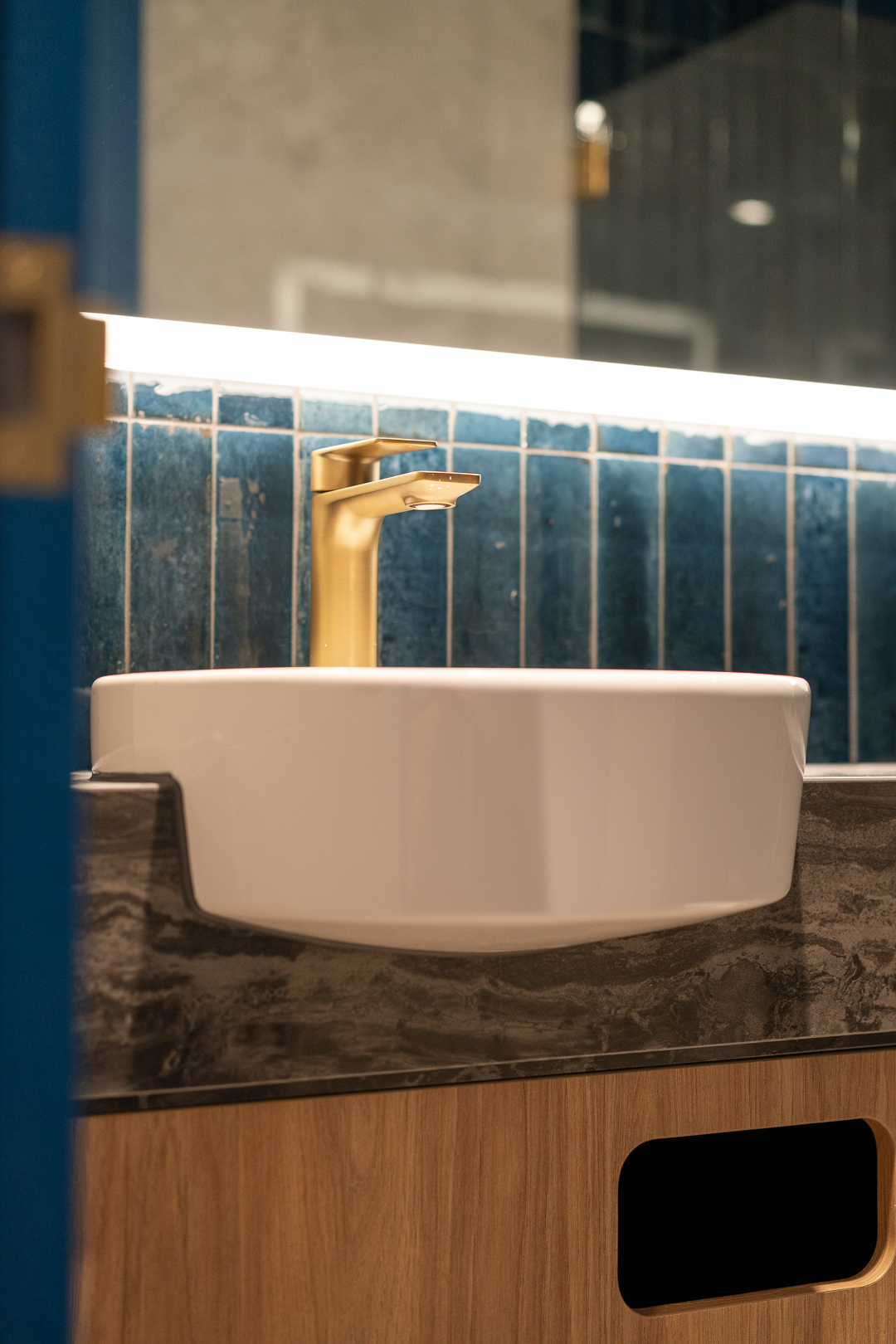 Modern bathroom interior featuring a white basin sink, gold faucet, and blue tiled backsplash with under-cabinet lighting.