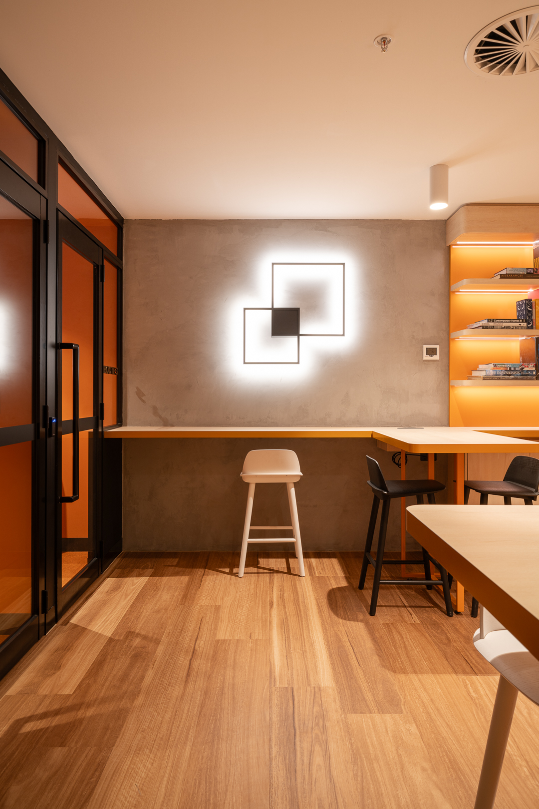 Modern workspace interior with wooden floors, a long desk with a stool, and a geometrically designed wall light in a brightly lit room.