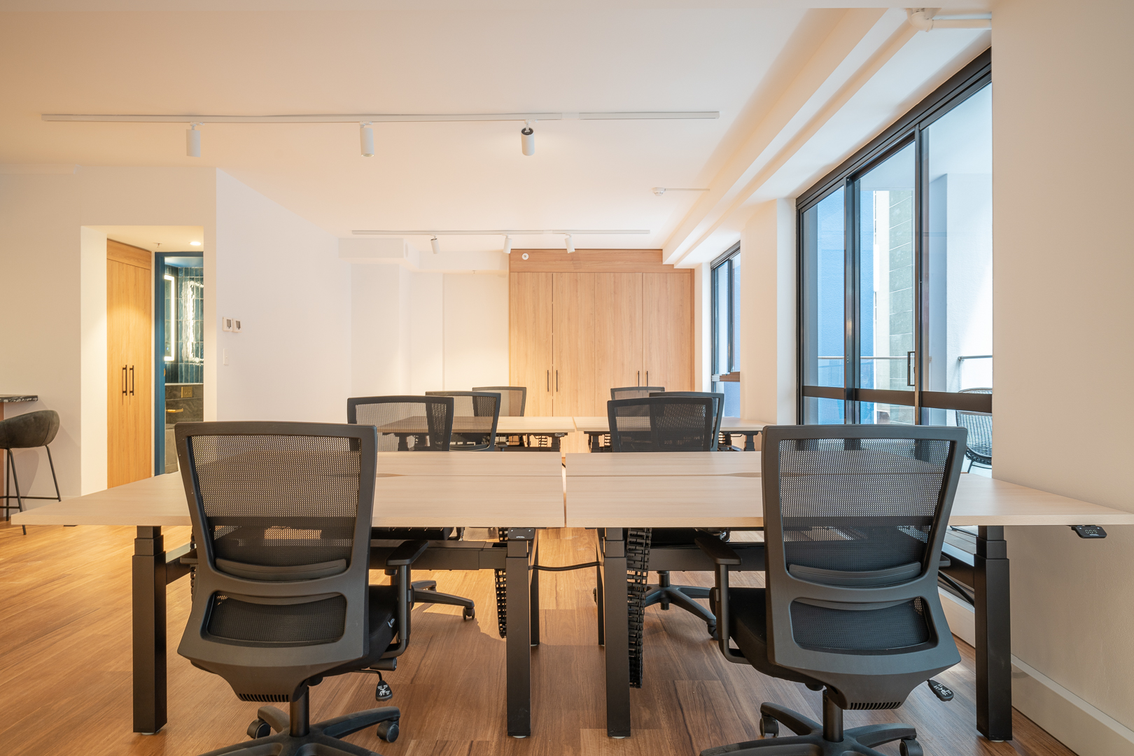 Office space with multiple desks and chairs in a brightly lit, modern interior featuring wood floors and large windows.
