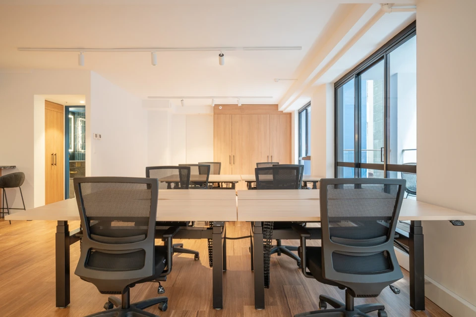 Office space with multiple desks and chairs in a brightly lit, modern interior featuring wood floors and large windows.