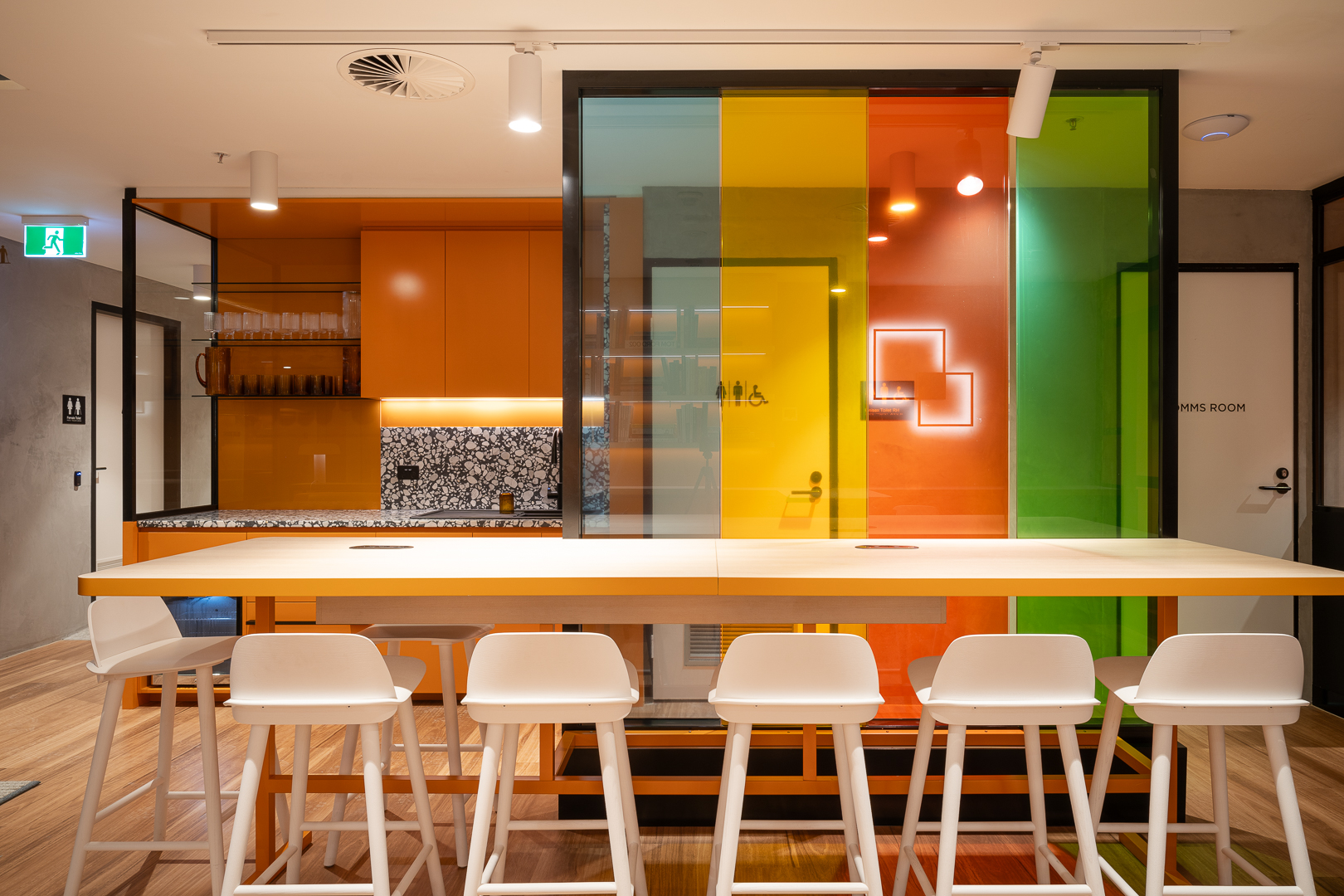 Interior view of a modern office space with a long communal table, white chairs, and colorful glass partitions in the background.