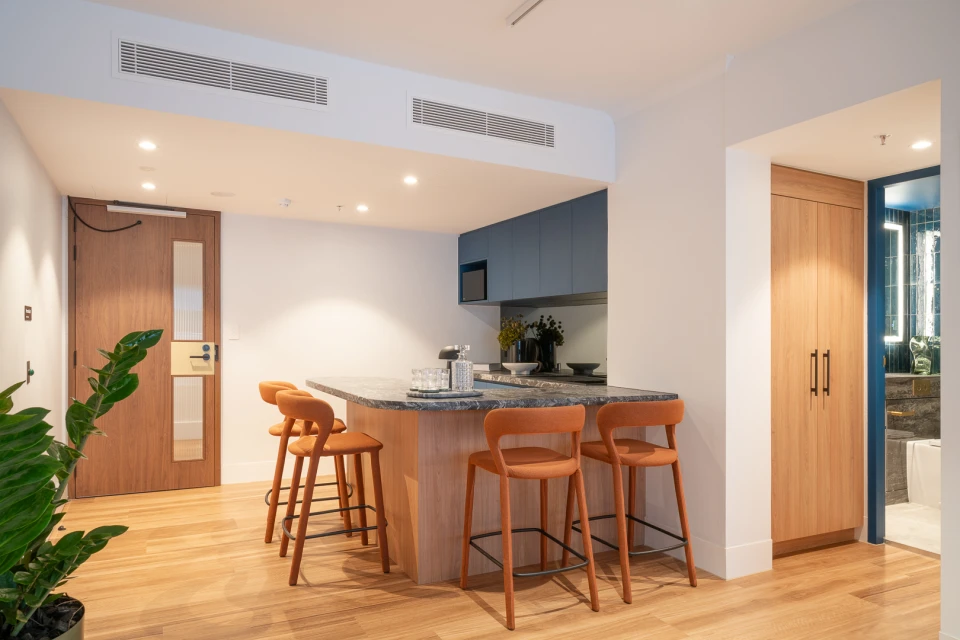 Interior view of an apartment studio kitchen with orange bar stools, a kitchen island, and light wood flooring and cabinetry.