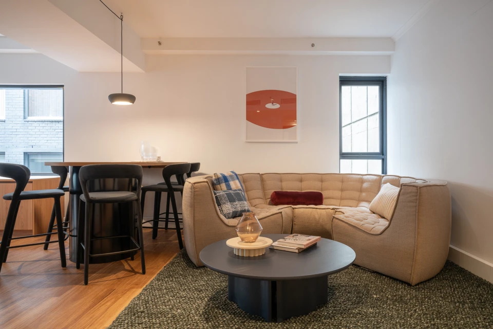 Interior view of a modern living room with a beige sectional sofa, round coffee table, black bar stools, and an abstract art print.