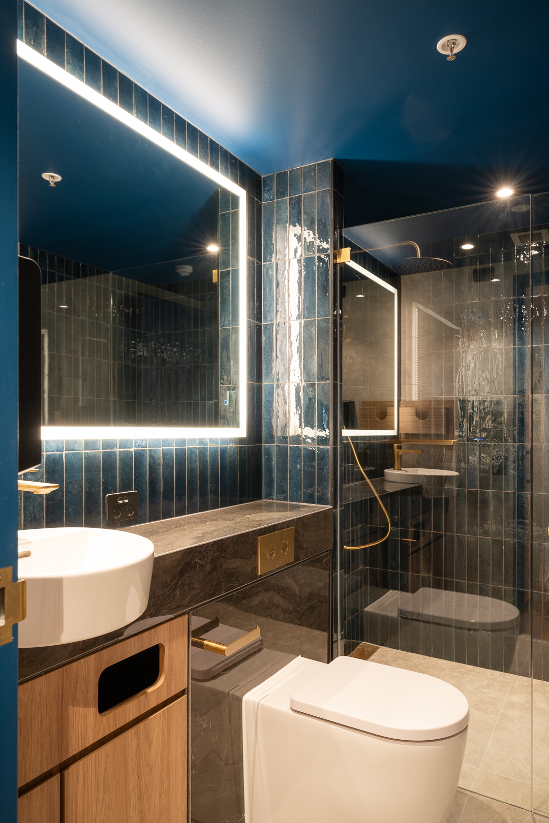 Modern bathroom interior featuring blue tiled walls, a bright LED mirror, white ceramic sink and toilet, wood cabinetry, and glass shower.