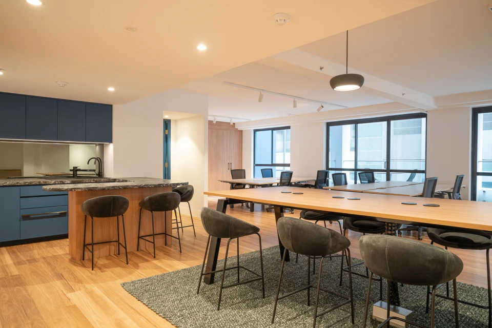 Contemporary office space featuring a kitchen area with bar stools and a large conference table with black chairs under bright lighting.