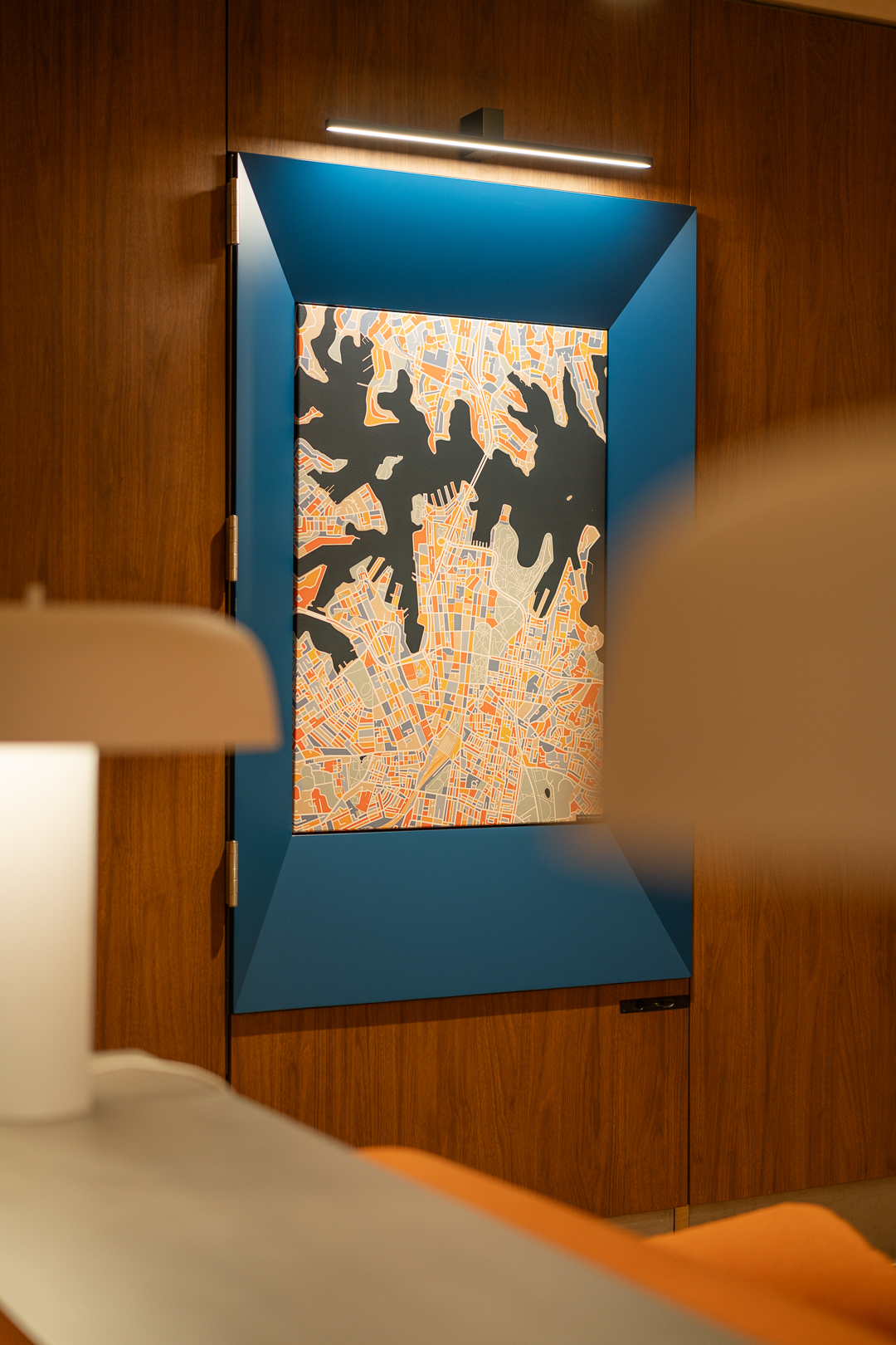 Abstract art with a blue frame hangs on a wooden wall, illuminated by a light fixture, complemented by a lamp and bed in the foreground.