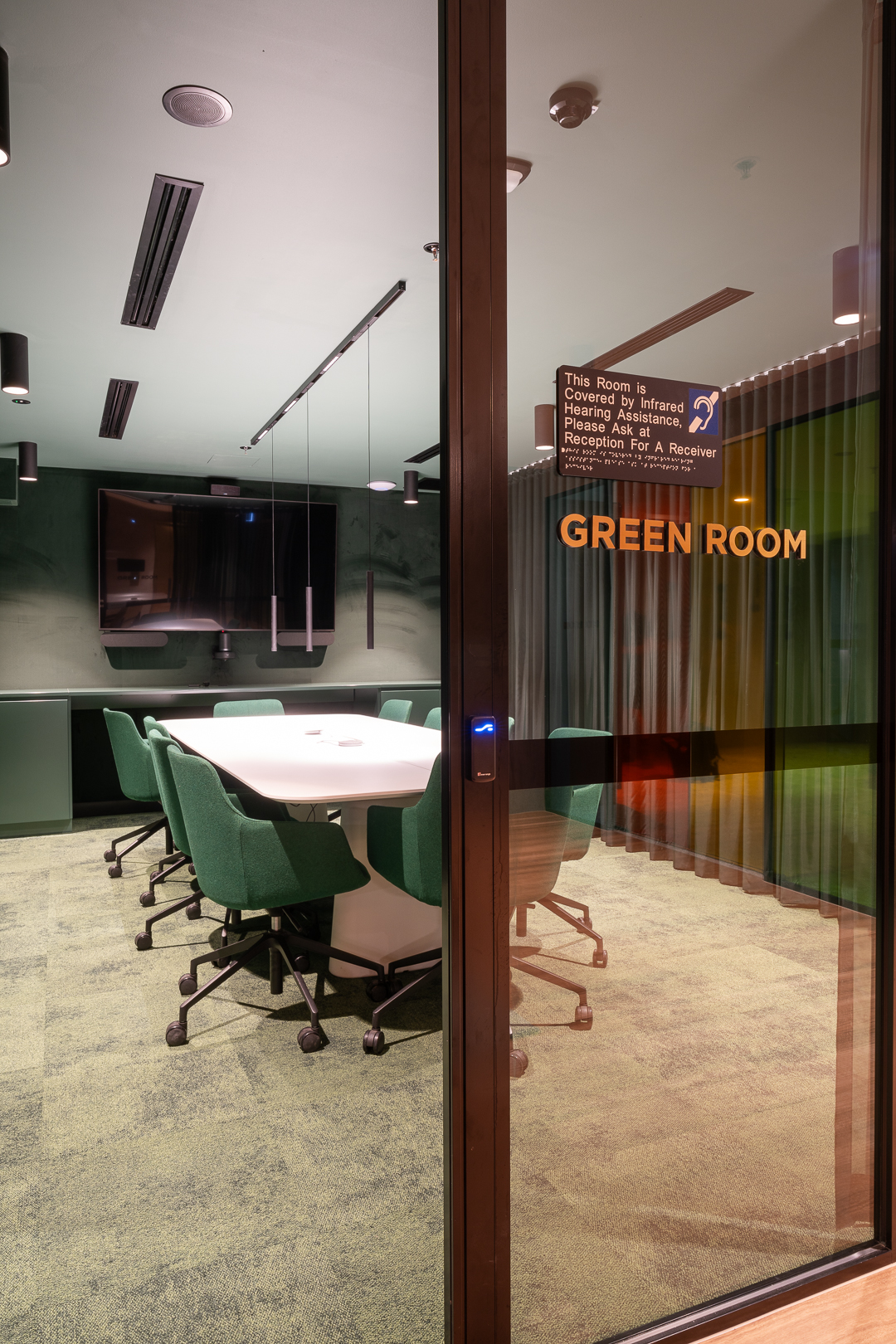 Conference room with a white conference table and green chairs viewed through a glass door with green walls in the background.