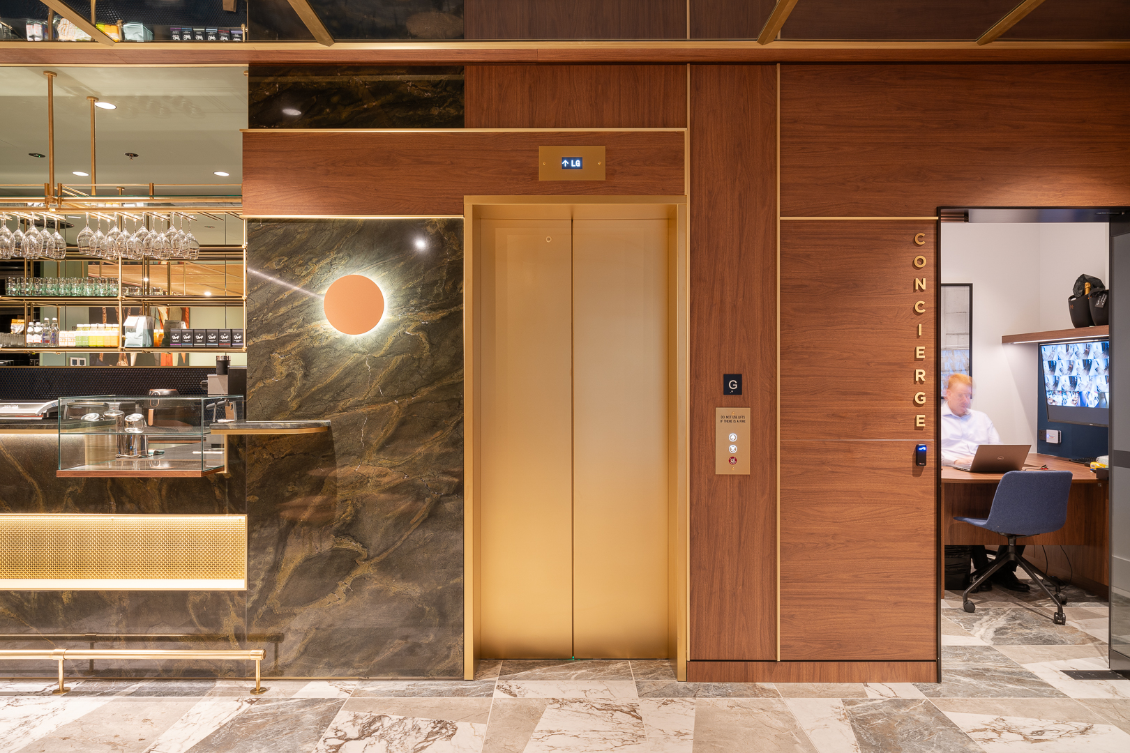 Interior of a hotel or office lobby featuring a golden elevator, marble walls, and a concierge desk with a person working on a laptop.