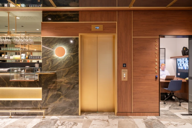 Interior of a hotel or office lobby featuring a golden elevator, marble walls, and a concierge desk with a person working on a laptop.