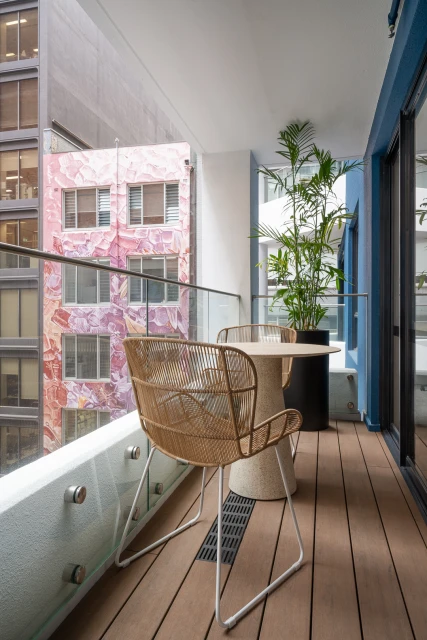 Indoor balcony with two wicker chairs, small round table, potted plant, and view of building with mural through glass railing.
