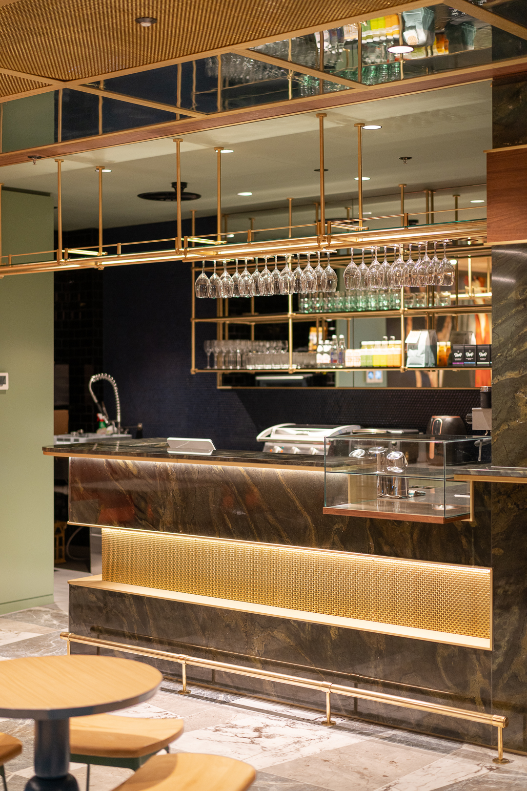 Interior view of a modern bar area featuring a counter with a marble top, geometric gold detailing, glass racks, and a tiled ceiling.